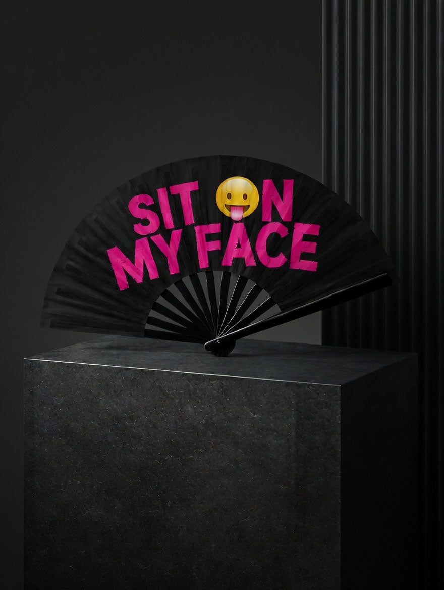 Black folding fan with pink text 'SIT ON MY FACE' and a yellow emoji on a dark background