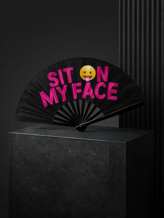 Black folding fan with pink text 'SIT ON MY FACE' and a yellow emoji on a dark background